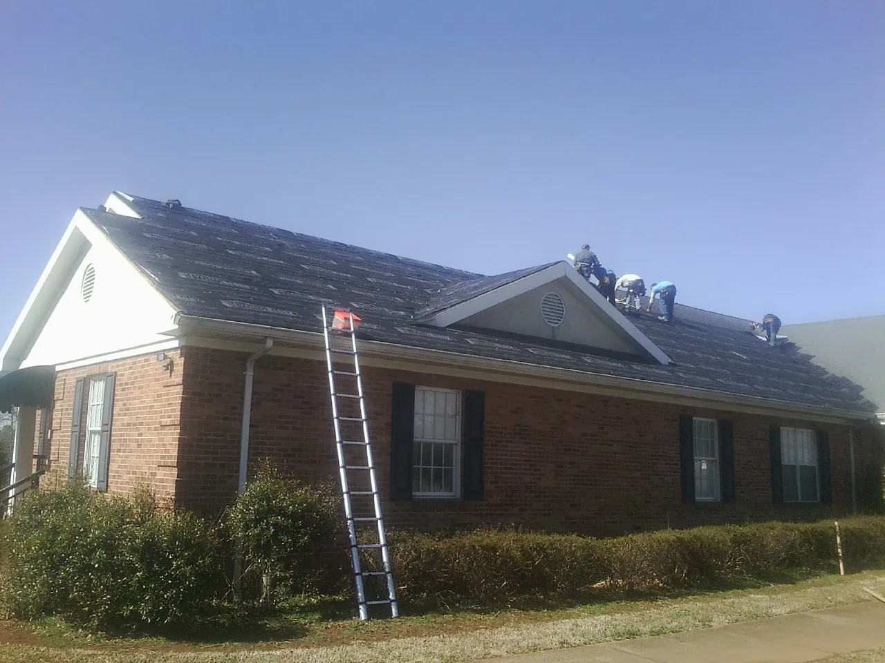 Crew tearing shingles off a brick ranch roof with a ladder set — Metal Roof Repair work by our crew in Mannsville