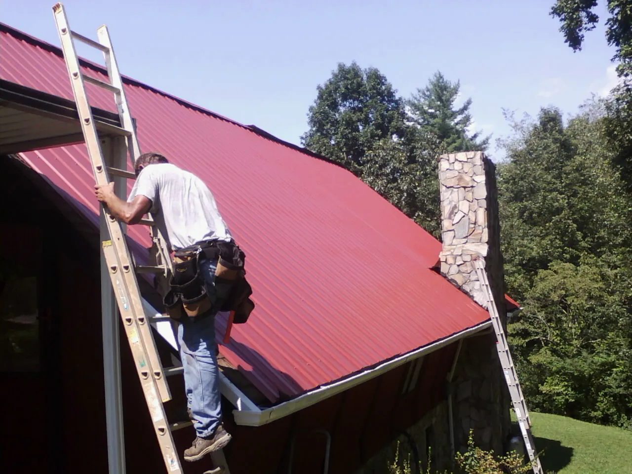 Roofer on a ladder beside a red metal roof and stone chimney — Soffit & Fascia Repair by Roofing Brothers in Mannsville